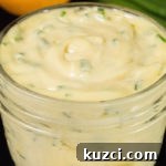 A small jar of lemon chive mayo sits on a black table with chives and a lemon in the background