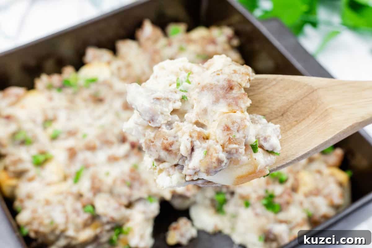 A wooden spoon holds a serving of biscuits and gravy with sausage, over a baking dish filled with the same dish, garnished with chopped herbs.