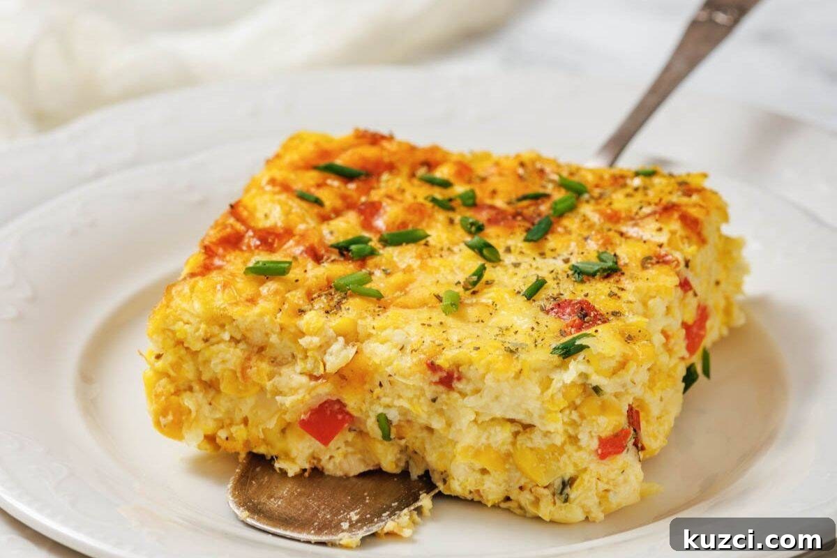 A square slice of corn pudding casserole with eggs, red bell pepper, and chives sits on a white plate with a serving spoon underneath.