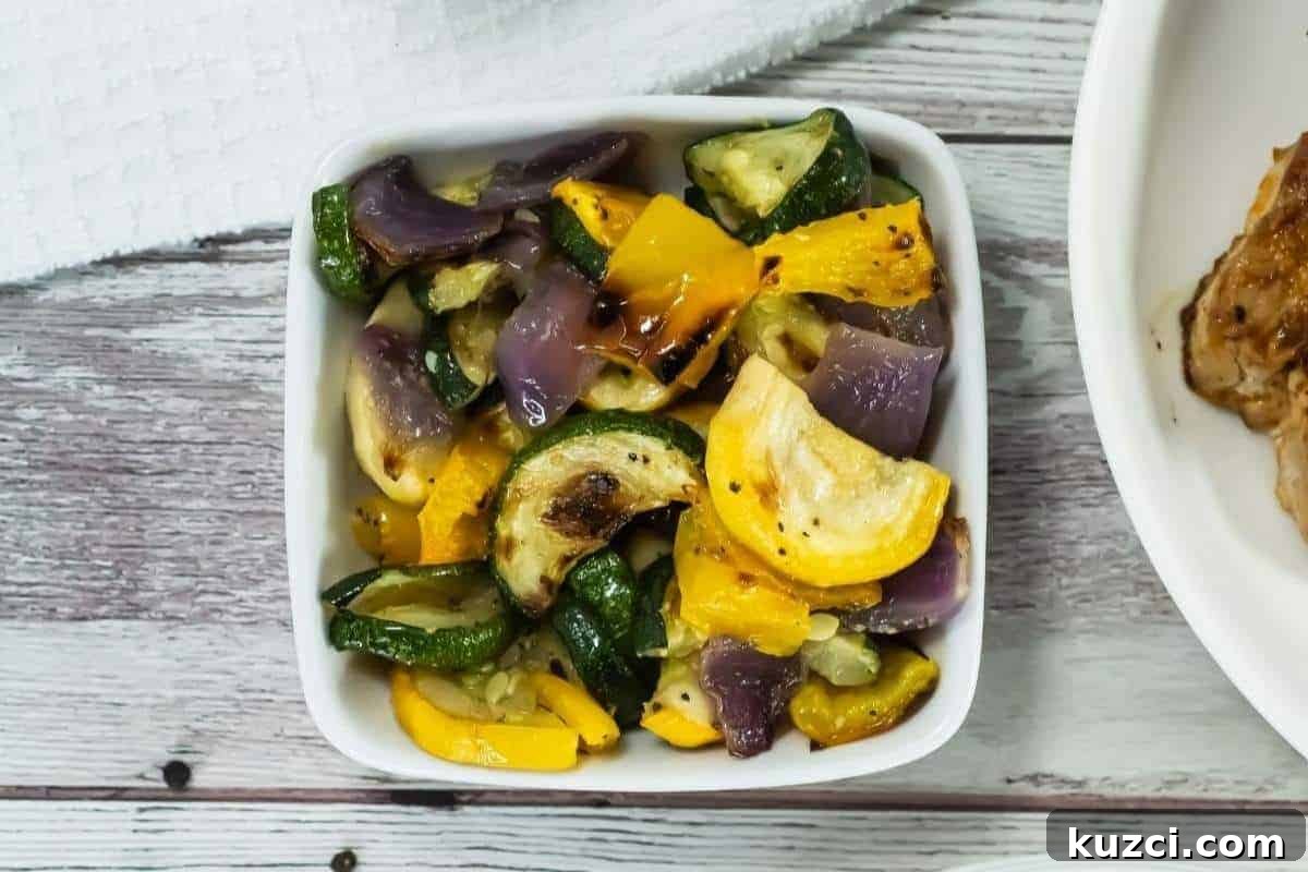 A white bowl filled with broiled vegetables, including zucchini, yellow squash, and red onion, sits on a wooden surface next to a white towel.