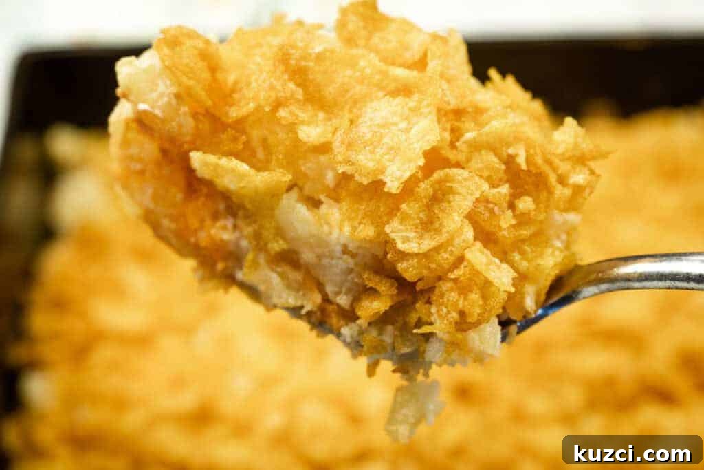 A close-up of a spoonful of Funeral potatoes with Cream Cheese, topped with crushed cornflakes, with the rest of the casserole in the background.