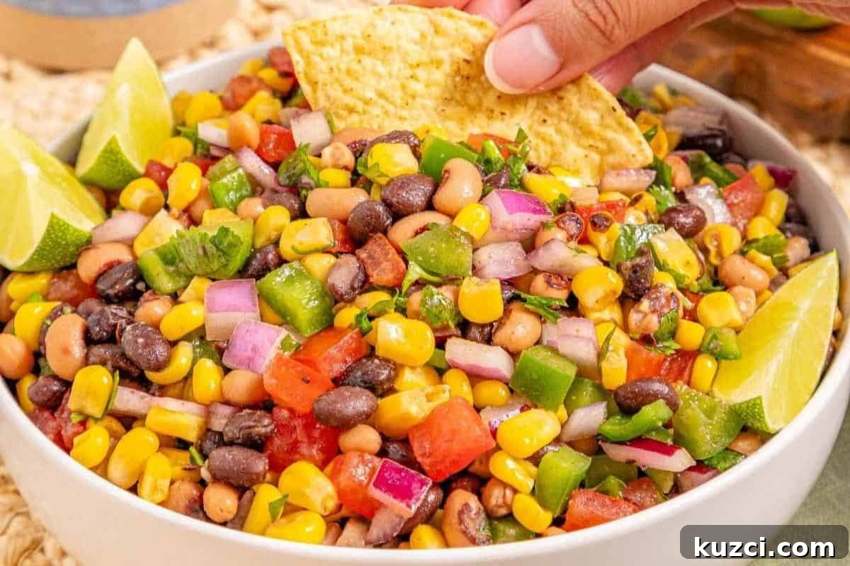A bowl of Cowboy Caviar, garnished with lime wedges; a hand dips a tortilla chip into the salad.