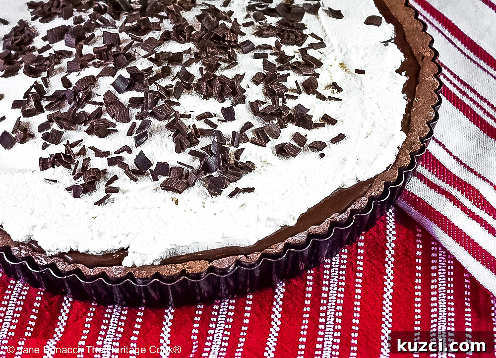 Chocolate Mocha Tart the whole tart, with the chocolate filling covered with a whipped topping and sprinkled with shaved chocolate pieces, some with a slice on a plate in front of the rest of the tart on red and white cloths © 2025 Jane Bonacci, The Heritage Cook.