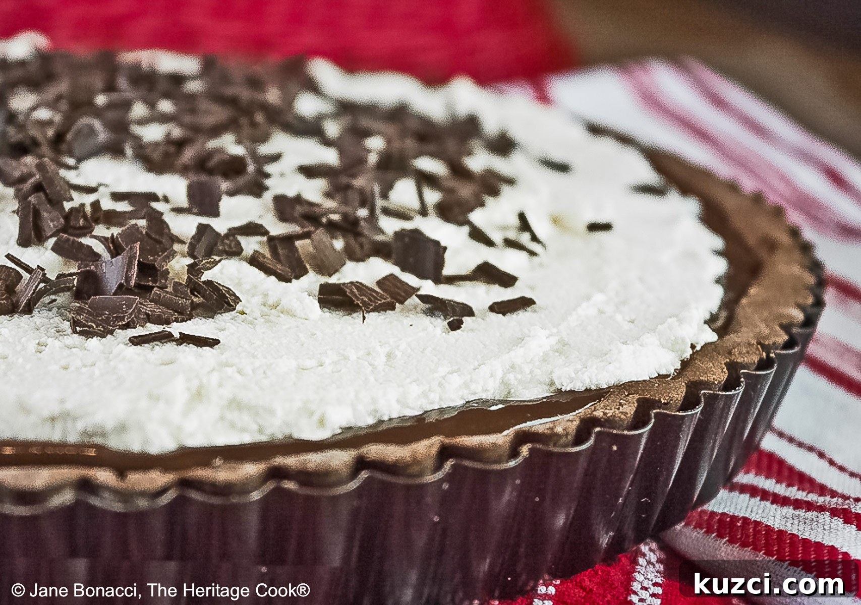 Chocolate Mocha Tart the whole tart, with the chocolate filling covered with a whipped topping and sprinkled with shaved chocolate pieces, some with a slice on a plate in front of the rest of the tart on red and white cloths © 2025 Jane Bonacci, The Heritage Cook.