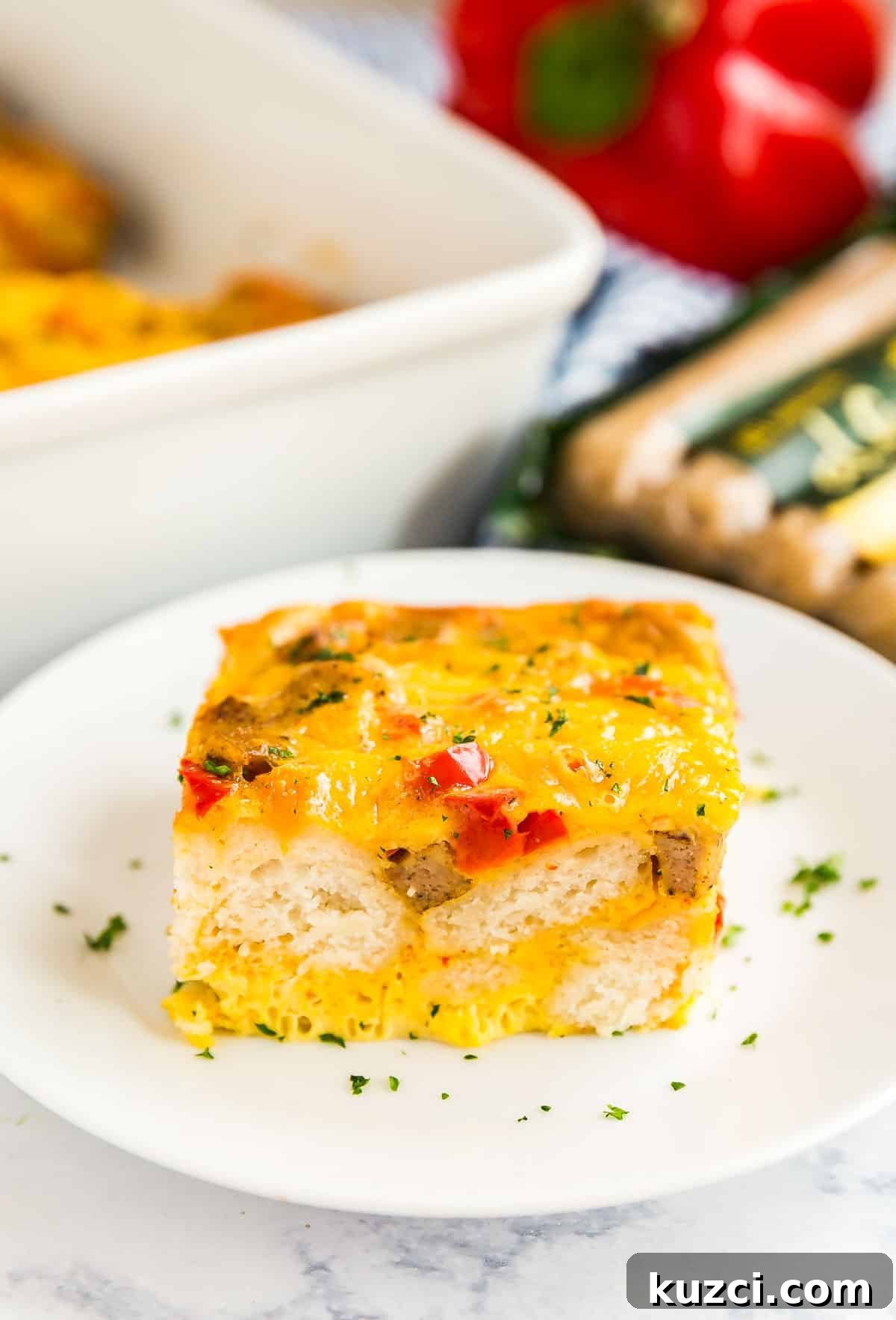 A piece of biscuit breakfast casserole on a white plate with the pan behind it and a blue and white striped napkin.