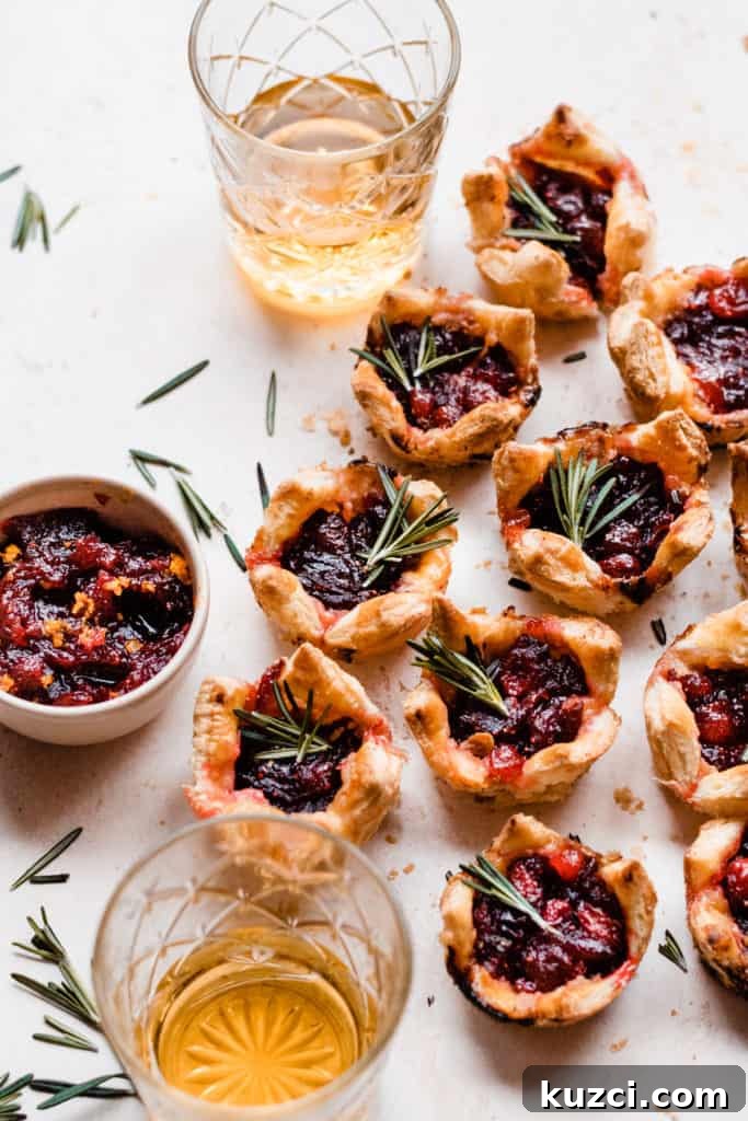 Gouda Puff Pastry Bites with Spiced Rum Cranberry Sauce 2 Puff pastry bites on a light surface surrounded by rosemary sprigs, glasses of rum, and a bowl of cranberry sauce