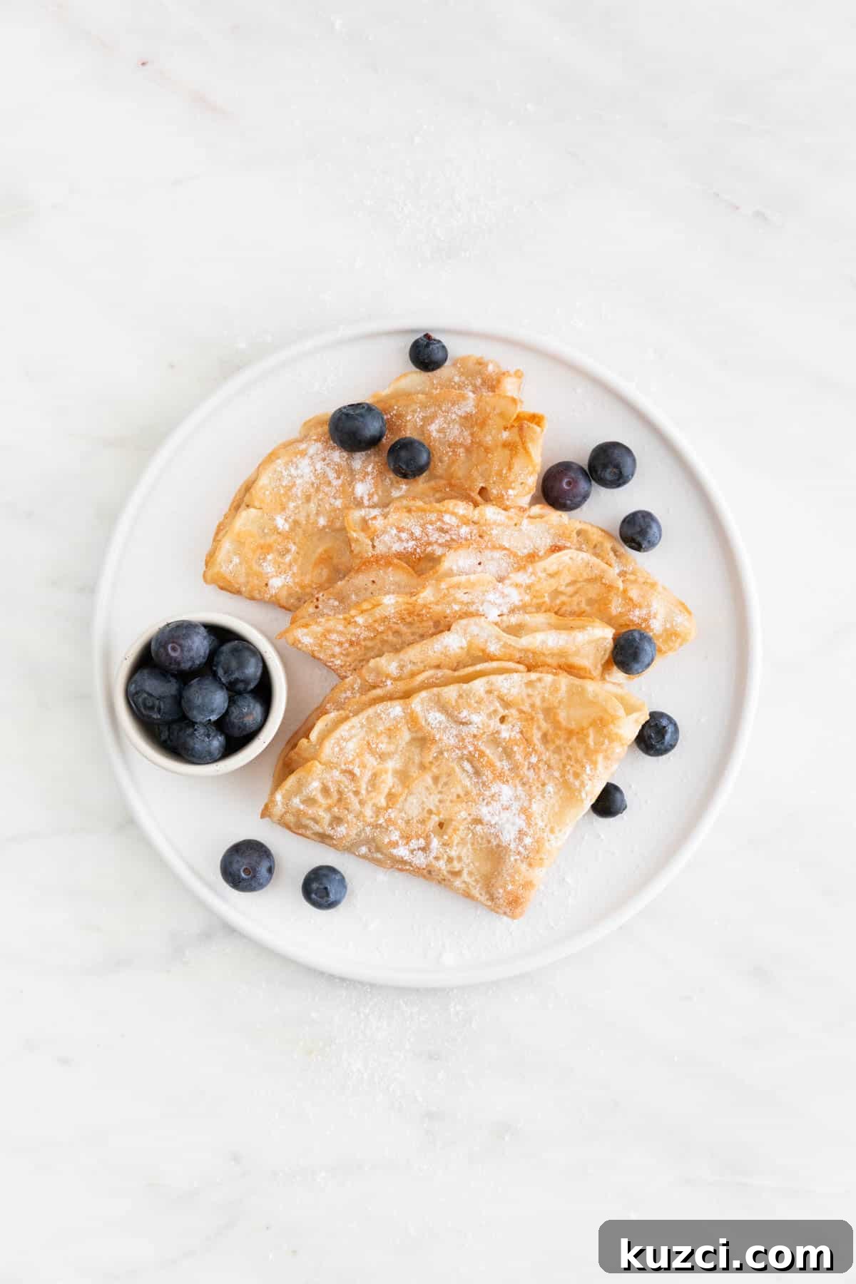 Overview of a plate with several folded vegan crepes topped with fresh blueberries and powdered sugar.
