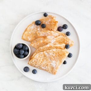 Overview of a plate with several folded vegan crepes topped with fresh blueberries and powdered sugar.