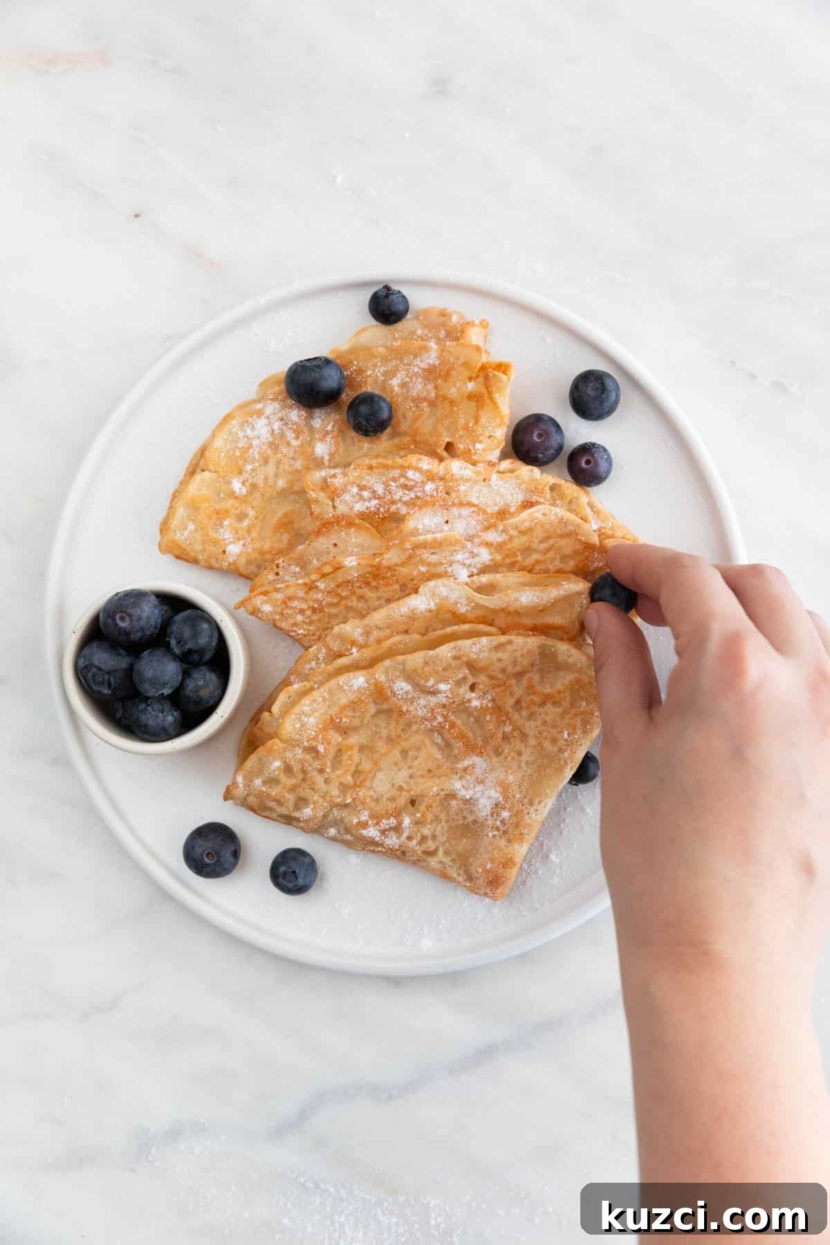 A hand adding fresh blueberries onto a plate with folded vegan crepes topped with powdered sugar.