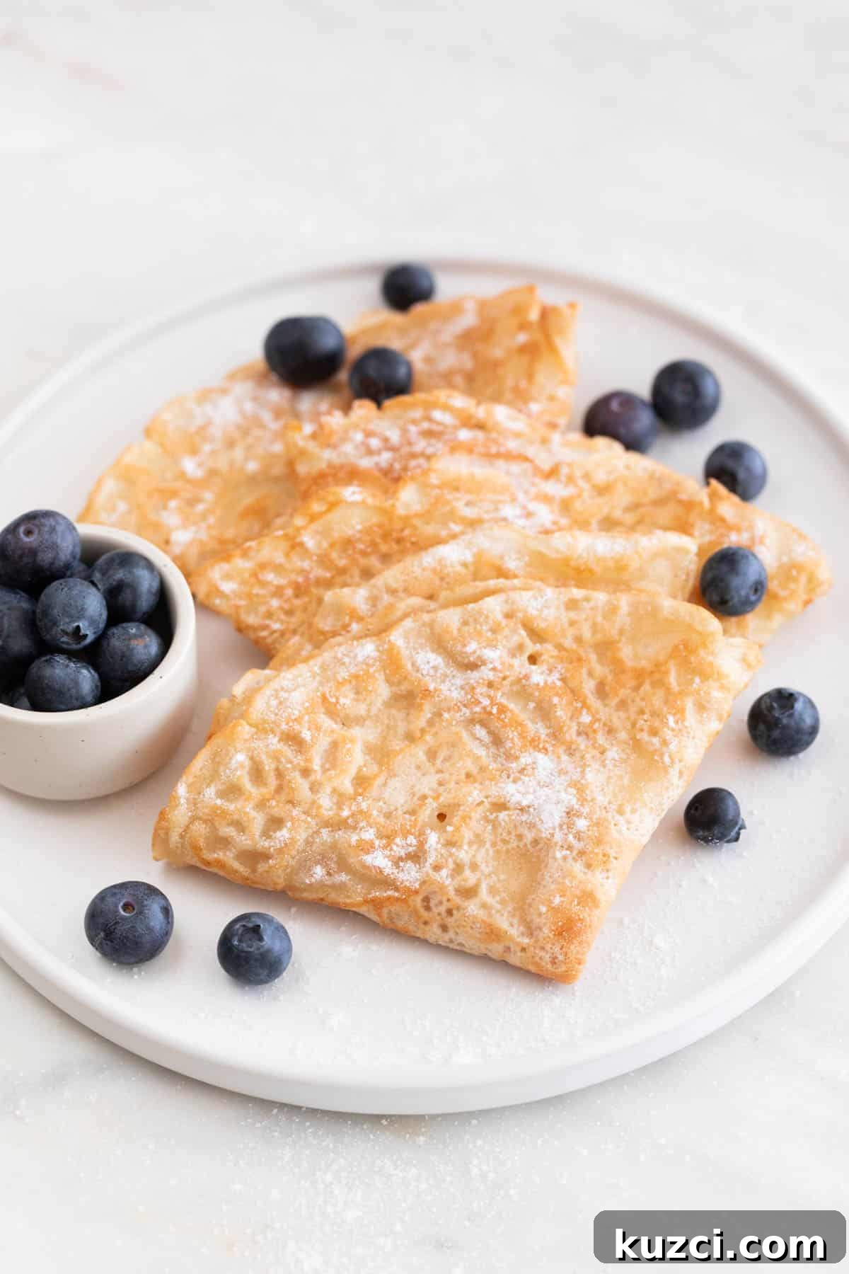 A plate with several folded vegan crepes with some blueberries and powdered sugar on top.