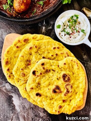 Birdsview of three sourdough naan placed on a wooden board and a white ceramic serving dish holding yoghurt with fresh herbs, chilli flakes and salt sprinkled over