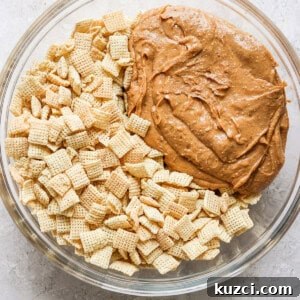 Peanut Butter Chex Bars Recipe 10 A glass bowl containing a mixture of cereal pieces and a large dollop of peanut butter on a light surface.