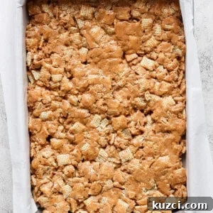 Peanut Butter Chex Bars Recipe 11 A baking tray filled with a peanut butter rice krispie treats mixture, lined with parchment paper on a light textured surface.