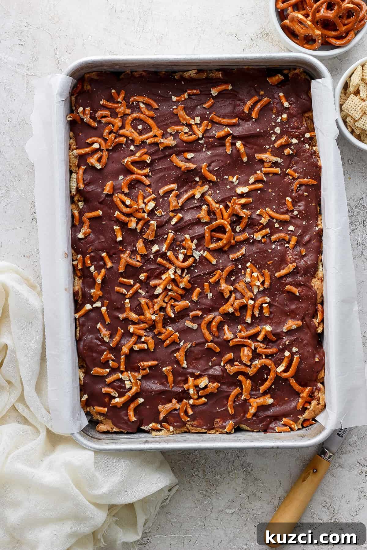 Peanut Butter Chex Bars Recipe 6 A tray of chocolate-covered toffee sprinkled with pretzel pieces on a kitchen counter, accompanied by a cloth and a spatula.