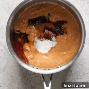 Peanut Butter Chex Bars Recipe 9 A stainless steel saucepan containing ingredients for a dish, including a reddish-brown sauce, visible spices, and a dollop of white substance, possibly coconut milk or cream, in the process of.