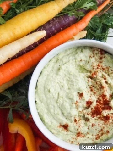 Edamame hummus in a white serving bowl and topped with paprika surrounded by tri-colored carrots and slices of bell pepper.