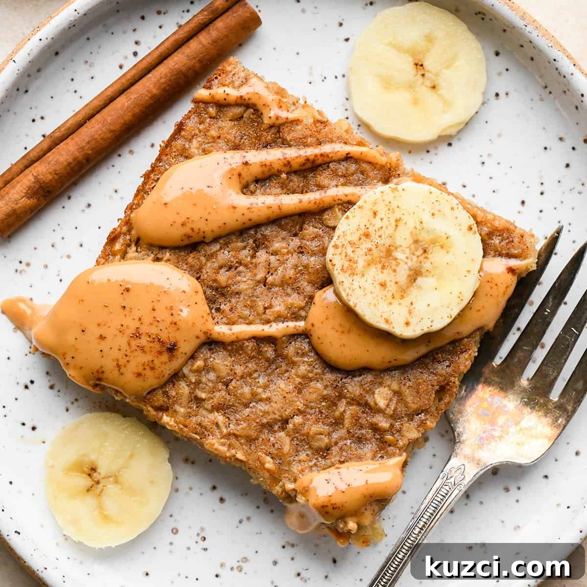 Peanut Butter Banana Baked Oatmeal a piece of Peanut Butter Banana Baked Oatmeal on a plate topped with a drizzle of peanut butter, bananas, and a sprinkle of cinnamon. There is a fork and a cinnamon stick on the plate too