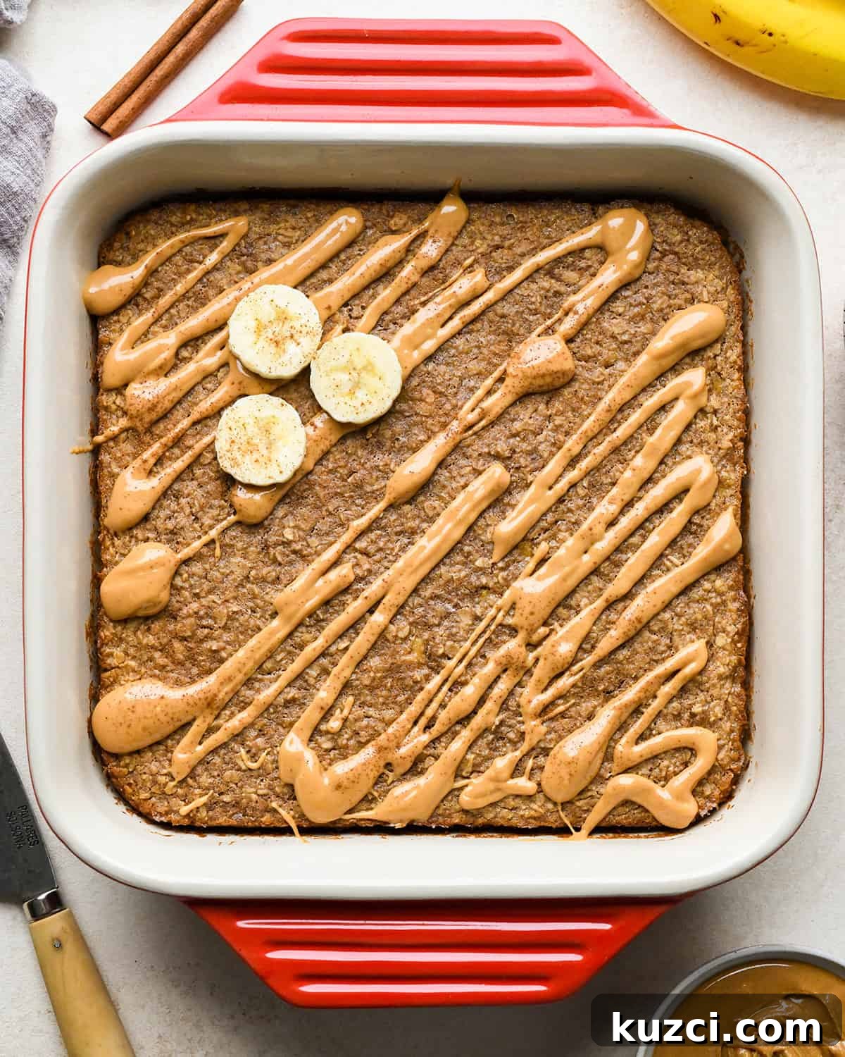 Peanut Butter Banana Baked Oatmeal Recipe 7 Peanut Butter Banana Baked Oatmeal in a red square baking dish drizzled with peanut butter, topped with 3 sliced bananas and cinnamon