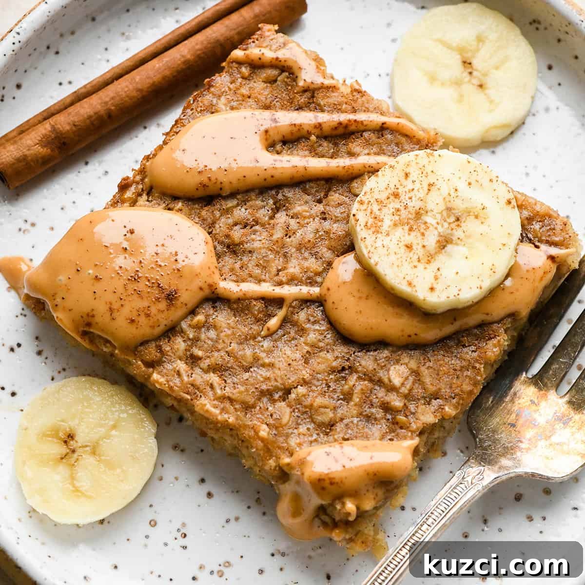 Peanut Butter Banana Baked Oatmeal Recipe 9 a square slice of Peanut Butter Banana Baked Oatmeal on a plate topped with a drizzle of peanut butter, sliced bananas, and a sprinkle of cinnamon. There is a fork and a cinnamon stick on the plate too
