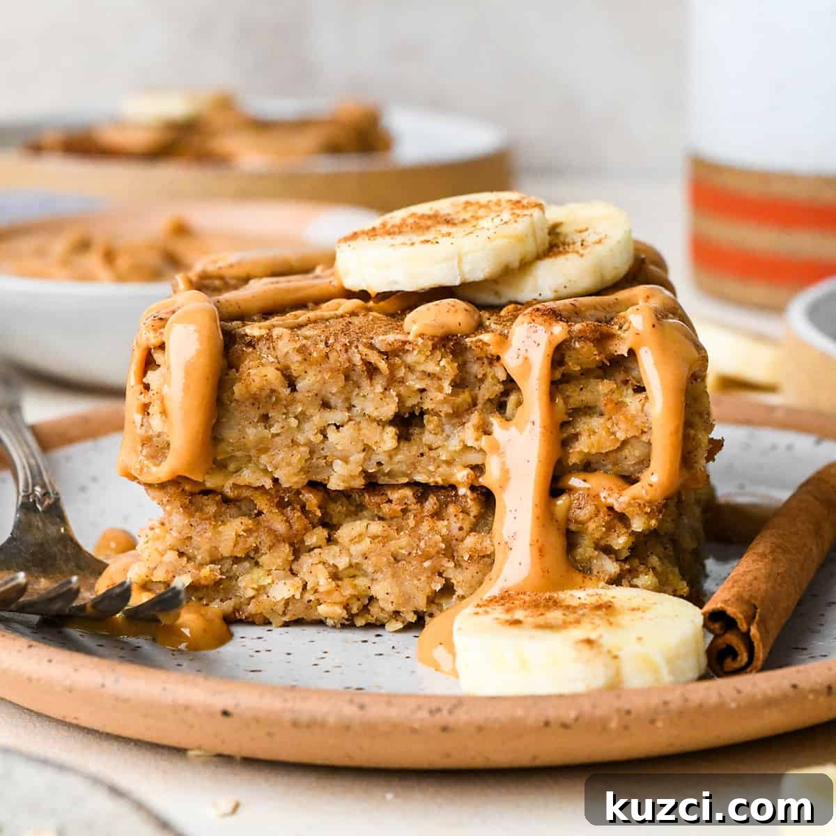 Peanut Butter Banana Baked Oatmeal Recipe 3 a stack of 2 slices of Peanut Butter Banana Baked Oatmeal topped with peanut butter that is melty and dripping down the sides, sliced bananas and a sprinkle of cinnamon. There is banana, a fork and a cinnamon stick on the plate and more bowls of oatmeal in the backgorund