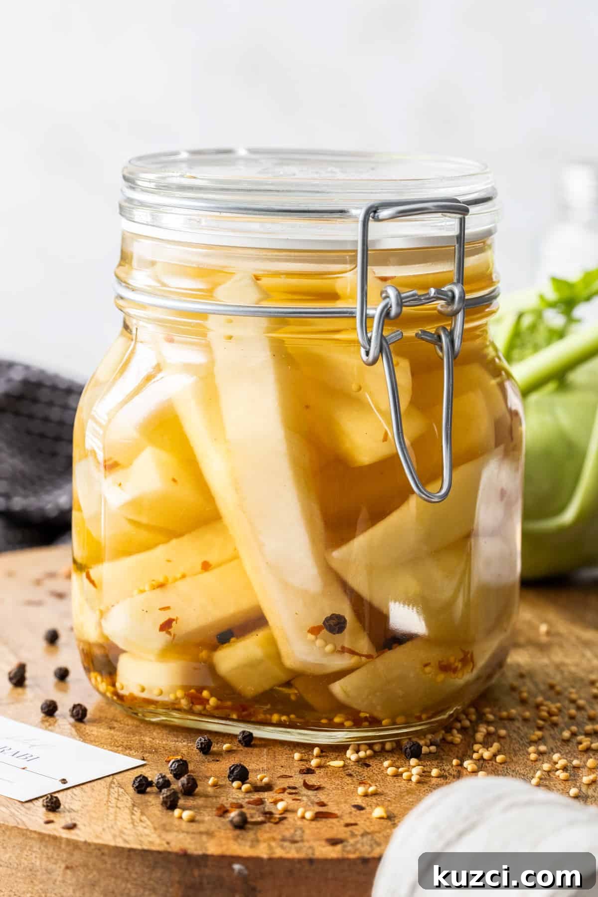 Jar of Pickled Kohlrabi, sitting on a round wooden board, with some herbs and spices around the edge.