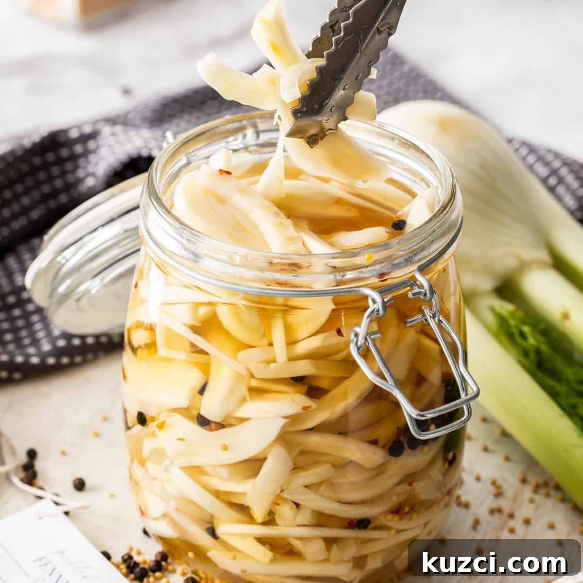 Jar of Pickled Fennel, sitting on a wooden board, with some tongs reaching in to pick up some pieces of fennel.