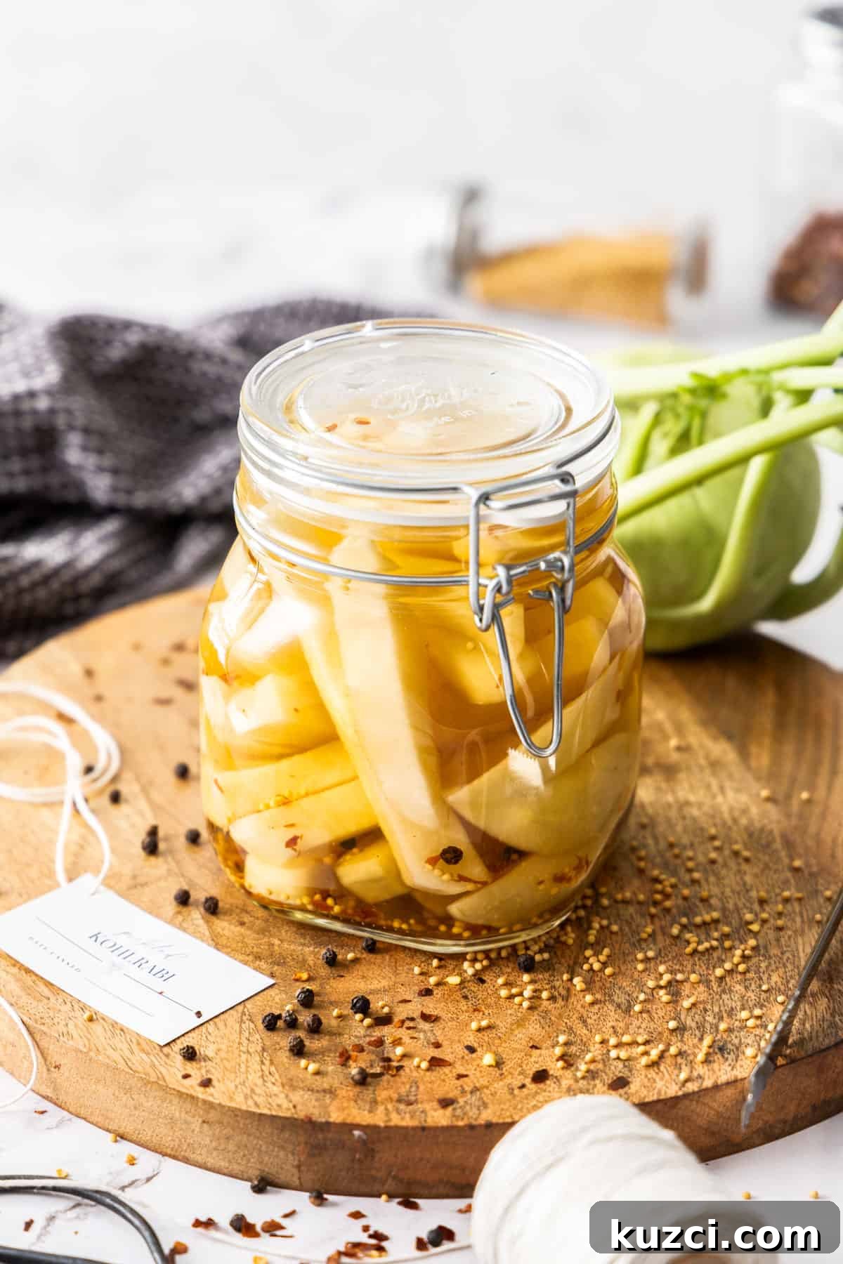 Jar of Pickled Kohlrabi, sitting on a round wooden board, with some herbs and spices around the edge.