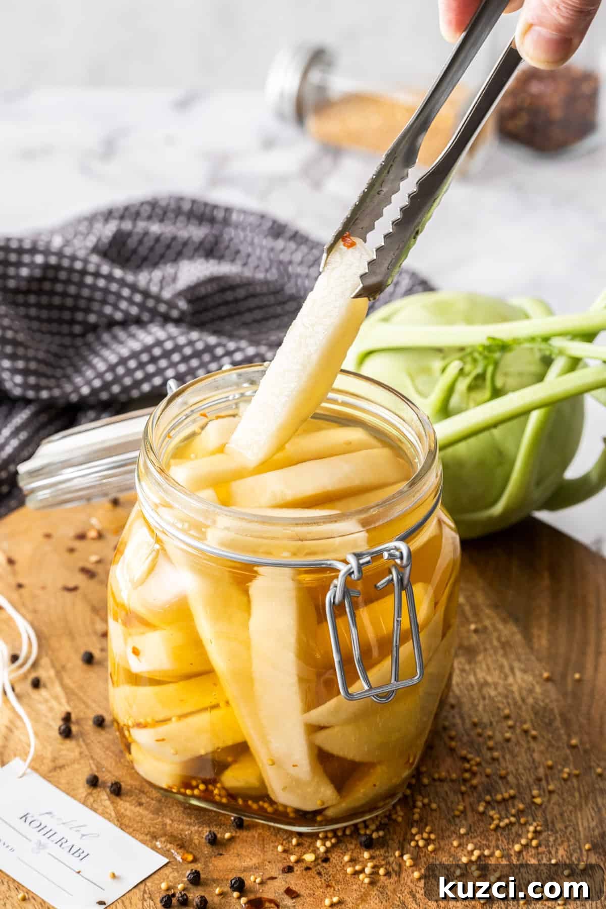 Jar of Pickled Kohlrabi, sitting on a round wooden board, with some tongs reaching in to pick up a piece of kohlrabi.