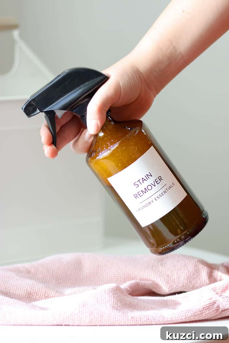 a glass amber spray bottle with a label that says 'stain remover laundry essentials' on it being sprayed at a stained baby outit