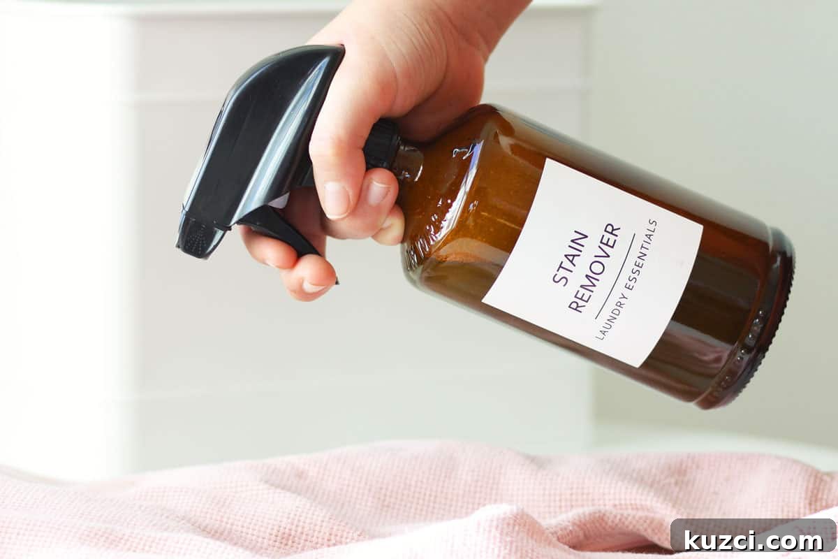 a glass amber spray bottle with a label that says 'stain remover laundry essentials' on it being sprayed at a stained baby outit