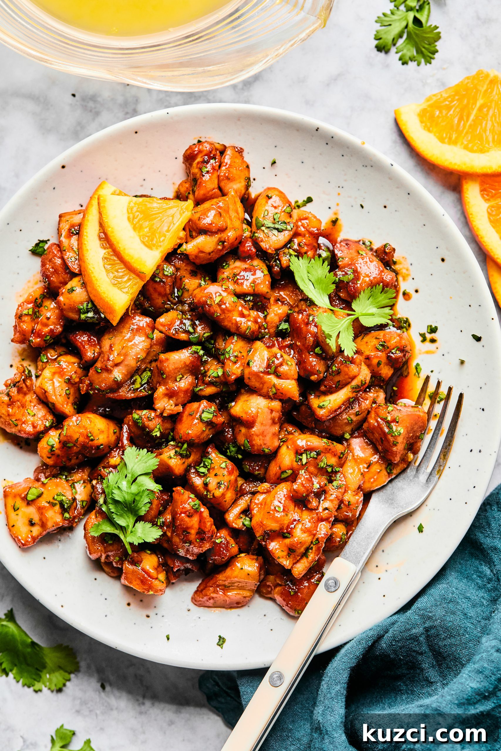 A small white serving plate filled with cilantro orange chicken bites sitting next to fresh orange slices and a small jar of orange juice