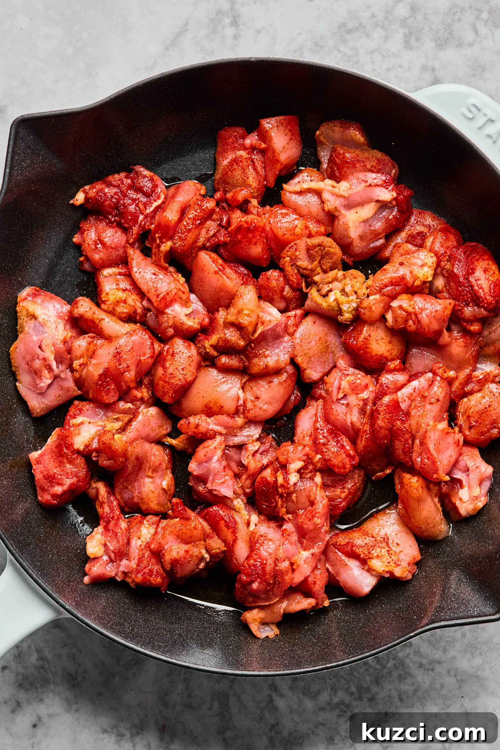 A large saute pan filled with raw chicken bites tossed in smoked paprika, cumin and salt.