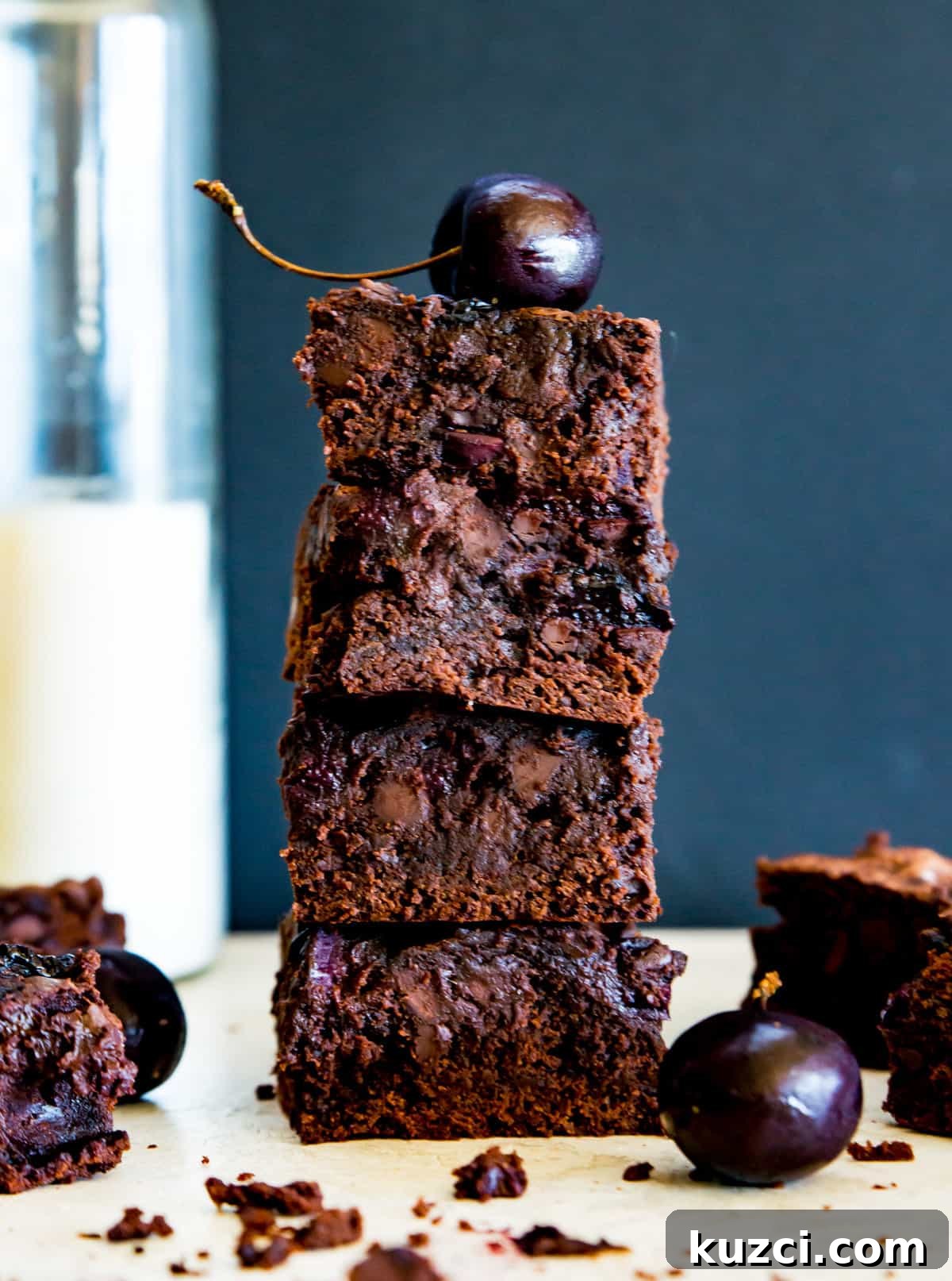 Fudgy Cherry Brownies with Dark Chocolate 9 A stack of chocolate cherry brownies with a cherry on top and a glass of milk behind them.