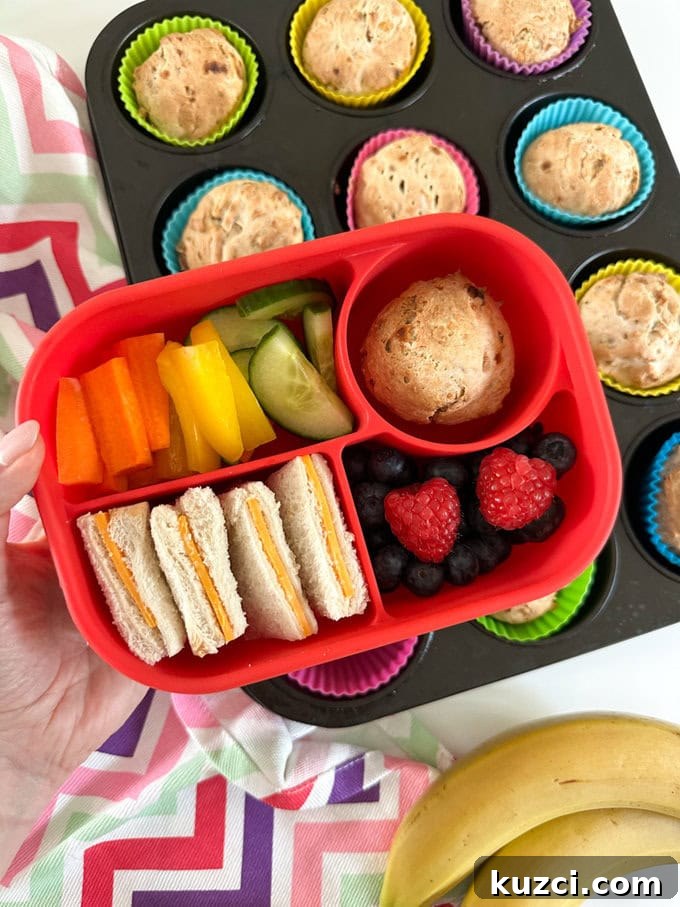 3 Ingredient Banana Muffins presented in a bright red Bento style lunchbox, with cheese sandwiches and fresh berries, cucumber slices and chopped yellow pepper and carrot's.