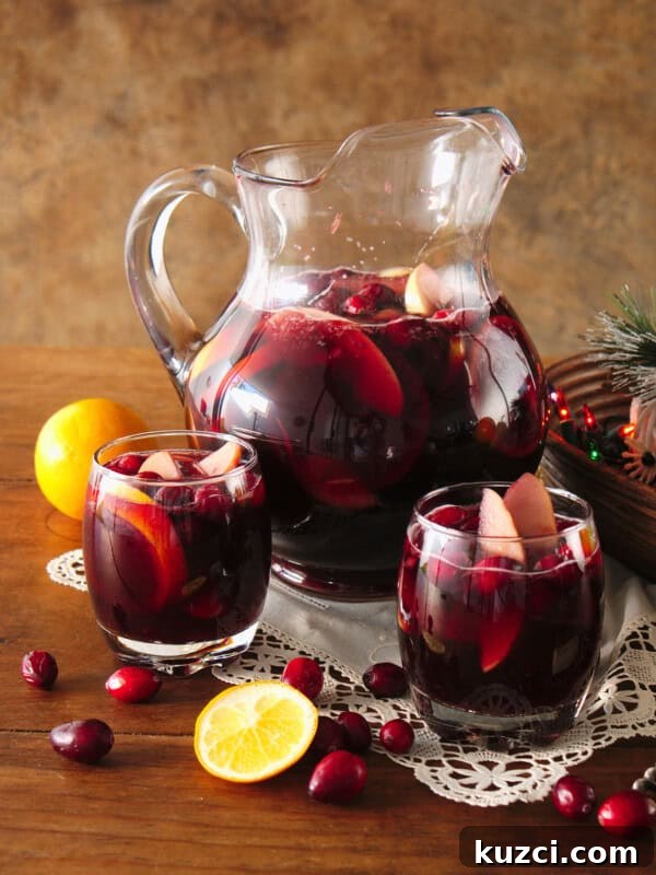 Witches' Brew Sangria: Spooky Halloween Cocktail 8 Clear glass pitcher and two rocks glasses filled with fruit-filled red Christmas sangria on a wooden table, styled with scattered cranberries, a white doily, and a bowl of multicolored Christmas lights.