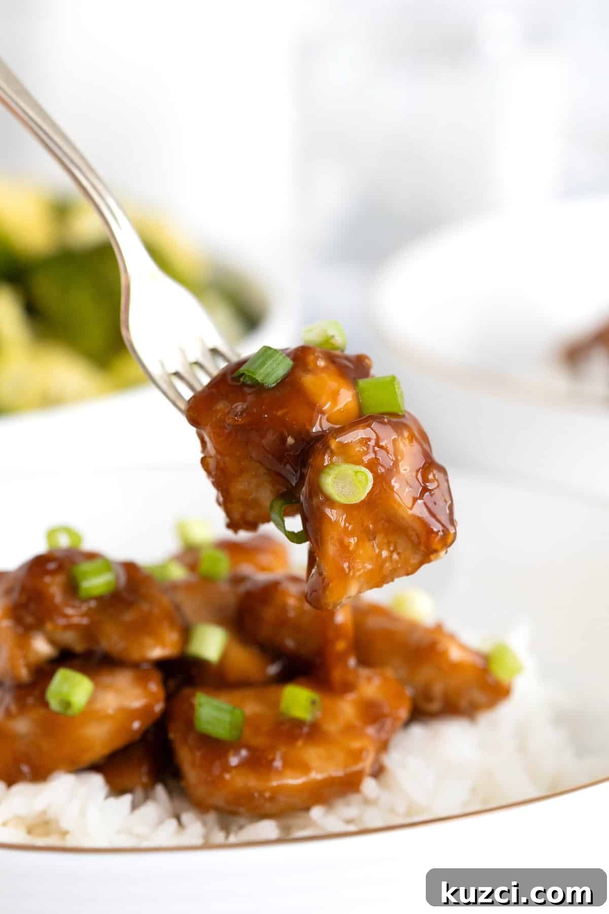 Two chunks of orange chicken sprinkled with green onion on a fork. A dish of rice and orange chicken is in the background.
