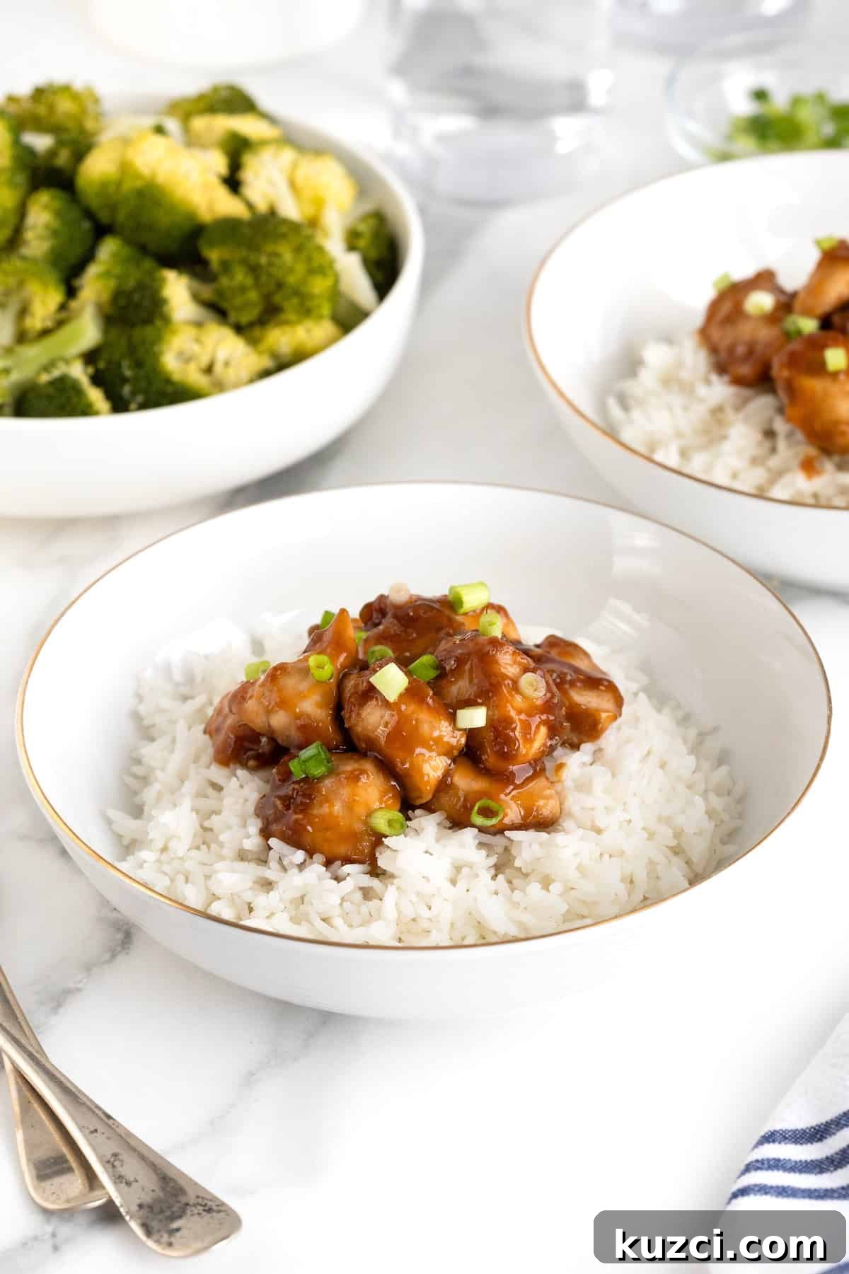 A white, gold-rimmed bowl of rice topped with orange chicken. A bowl of broccoli sits in the upper left of the photo.