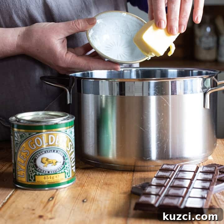 No-Bake Chocolate Cornflake Cakes Recipe 4 womans hands holding a stick of butter over a large silver saucepan on a wooden kitchen counter surrounded by baking mess