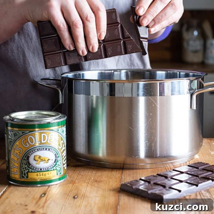 No-Bake Chocolate Cornflake Cakes Recipe 5 womans hands breaking dark chocolate pieces over a large silver saucepan on a wooden kitchen counter surrounded by baking mess