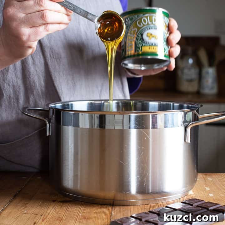 No-Bake Chocolate Cornflake Cakes Recipe 6 womans hands spooning golden syrup from a green can into a large silver saucepan on a wooden kitchen counter surrounded by baking mess
