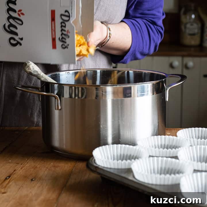 No-Bake Chocolate Cornflake Cakes Recipe 7 womans hands pouring yellow cornflakes from a white cereal box into a large silver saucepan on a wooden kitchen counter