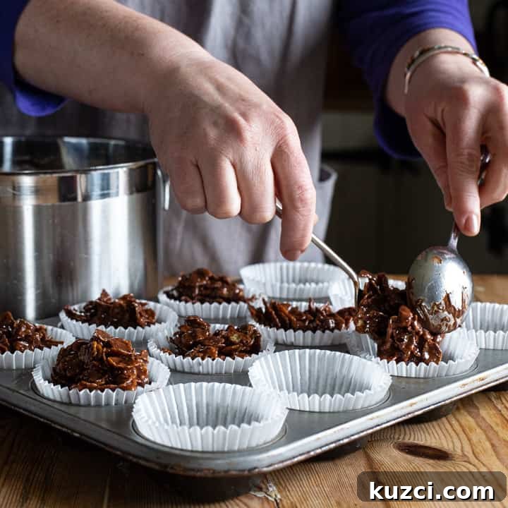 No-Bake Chocolate Cornflake Cakes Recipe 9 womans hands spooning chocolate covered cornflakes into white paper cupcake cases on a silver tray