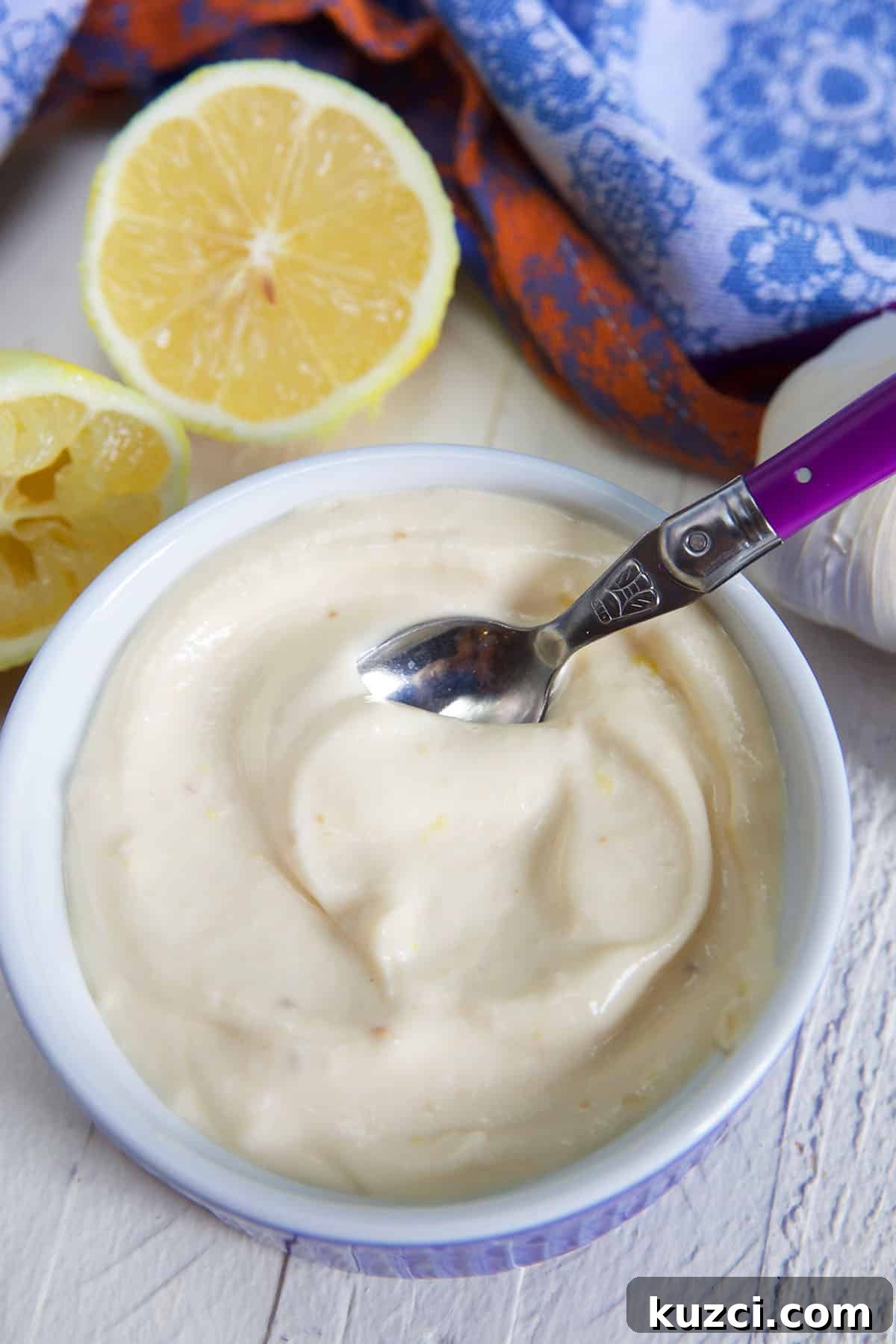 How to Make Roasted Garlic Aioli 4 garlic aioli in a blue and white ramekin with a purple handled spoon in it and a sliced lemon on the side.