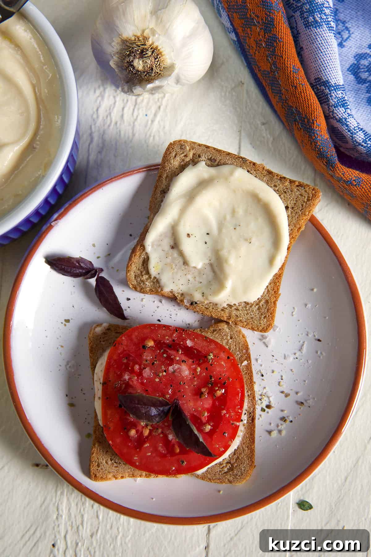 How to Make Roasted Garlic Aioli 5 Garlic Aioli on a piece of cocktail rye bread with a tomato slice on the side topped with purple basil leaves on a white plate.