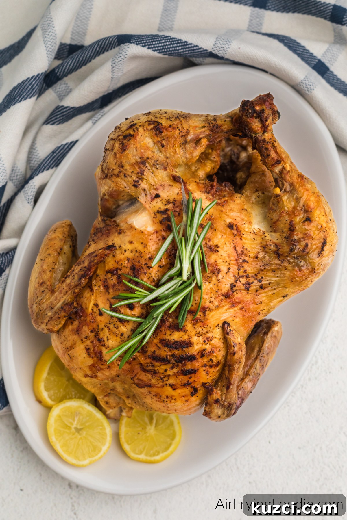 Crispy Whole Chicken in the Air Fryer 2 Air fried whole chicken on a white serving plate with sprigs of rosemary and lemon slices.