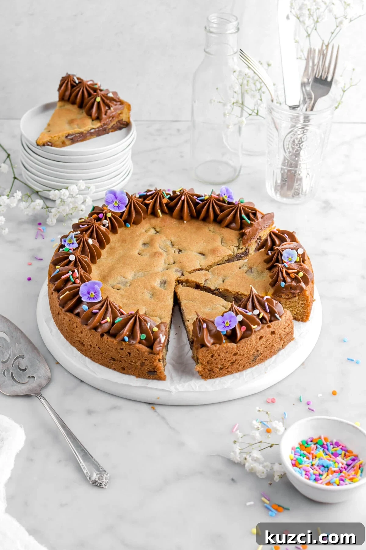 Chocolate Chip Cookie Cake with Chocolate Buttercream 2 angled shot of chocolate chip cookie cake with two slices cut into it on upside down plate with frosting piped around the edges, frosting, and purple flowers on top, bowl of sprinkles beside, stack of plates with another slice of cookie cake on top.