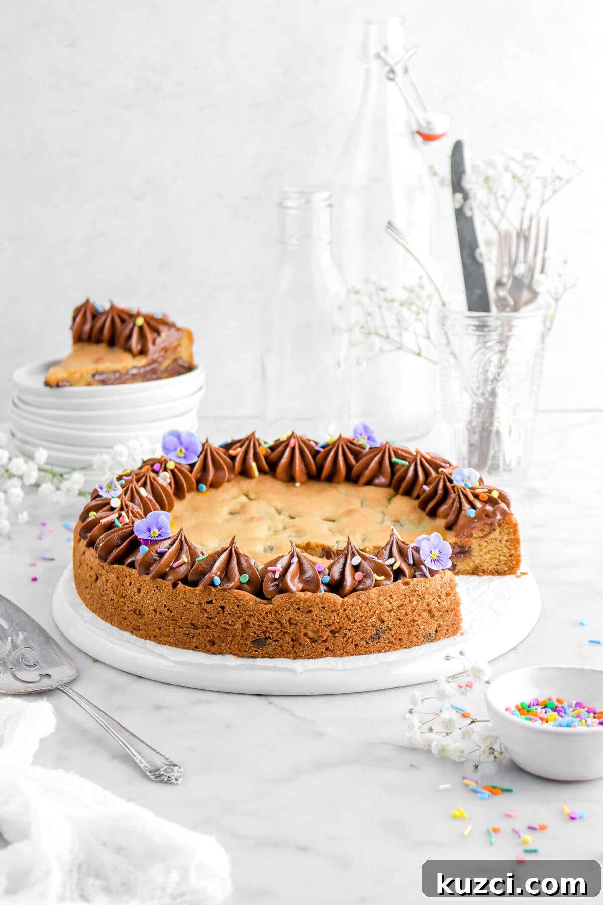 Chocolate Chip Cookie Cake with Chocolate Buttercream 24 cookie cake missing a slice on upside down white plate with bowl of sprinkles beside, a slice of cookie cake on stack of plates behind, with glass of utensils.