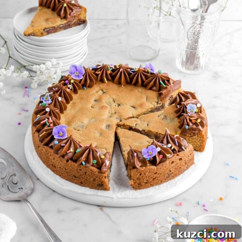 Chocolate Chip Cookie Cake with Chocolate Buttercream 27 angled shot of chocolate chip cookie cake with two slices cut into it on upside down plate with frosting piped around the edges, frosting, and purple flowers on top, bowl of sprinkles beside, stack of plates with another slice of cookie cake on top.