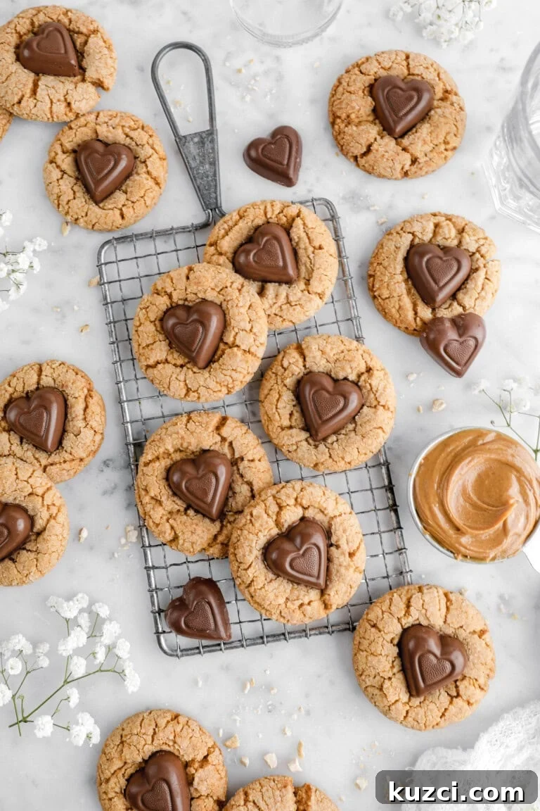 Peanut butter cookies with heart-shaped chocolate candies on top, arranged on a small wire cooling rack and a marble countertop. Some cookies are whole, while others are broken, revealing a soft interior. A small dish of peanut butter, scattered chocolate hearts, and delicate white flowers decorate the scene.