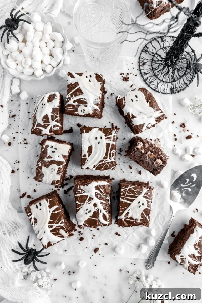cut marshmallow brownies on parchment paper with white chocolate ghosts on top, brownie crumbs around, and fake spiders.